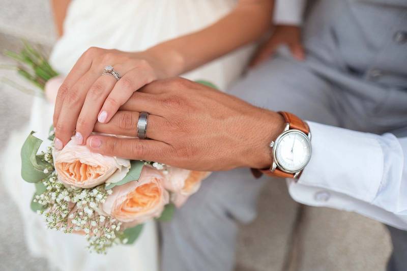 Quelle: pixabay.com/ Pexels, https://pixabay.com/de/photos/braut-paar-br%C3%A4utigam-h%C3%A4nde-1837148/ Braut und Bräutigam mit Ringen halten Hände auf Strauß